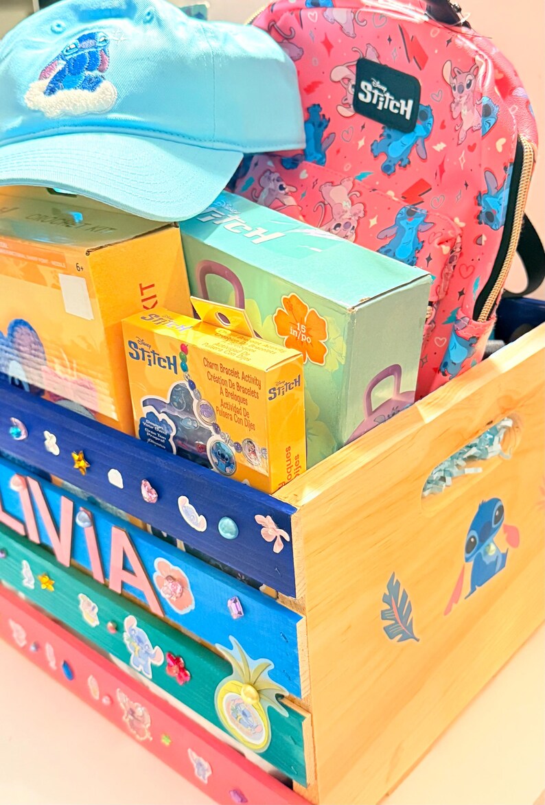 K&ouml;nnte beinhalten: Eine hellblaue Baseballkappe und ein pinkfarbener Rucksack mit der Figur Stitch sowie mehrere Kartons mit Stitch-Bastelsets befinden sich in einer Holzkiste. Die Kiste ist in verschiedenen Farben bemalt und mit Aufklebern und dem Namen "OLIVIA" verziert.