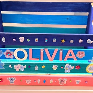 K&ouml;nnte beinhalten: Eine Holzkiste in Blau-, T&uuml;rkis- und Rosat&ouml;nen, verziert mit Aufklebern und dem Namen "OLIVIA" in rosa Buchstaben. Die Kiste hat eine rechteckige Form mit ausgeschnittenen Griffen.