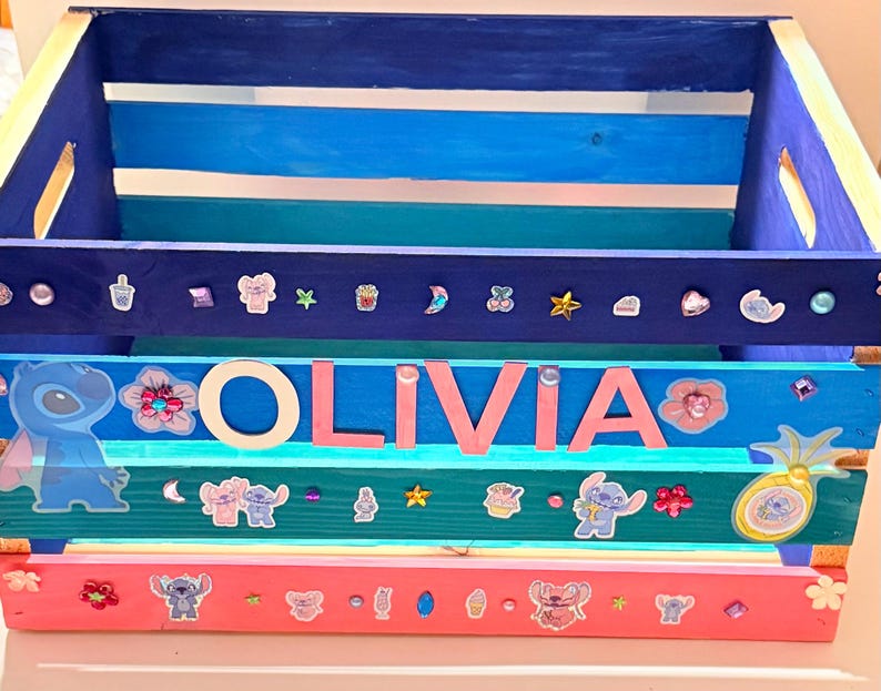 K&ouml;nnte beinhalten: Eine Holzkiste in Blau-, T&uuml;rkis- und Rosat&ouml;nen, verziert mit Aufklebern und dem Namen "OLIVIA" in rosa Buchstaben. Die Kiste hat eine rechteckige Form mit ausgeschnittenen Griffen.