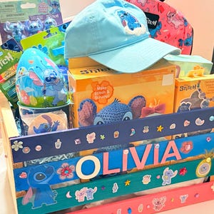 K&ouml;nnte beinhalten: Ein Geschenkkorb voller Stitch-Artikel. Enth&auml;lt eine hellblaue Baseballkappe mit einem gestickten Stitch-Design, einen rosa Rucksack, Spielzeug und eine Holzkiste, die mit dem Namen "OLIVIA" und Stitch-Aufklebern verziert ist.