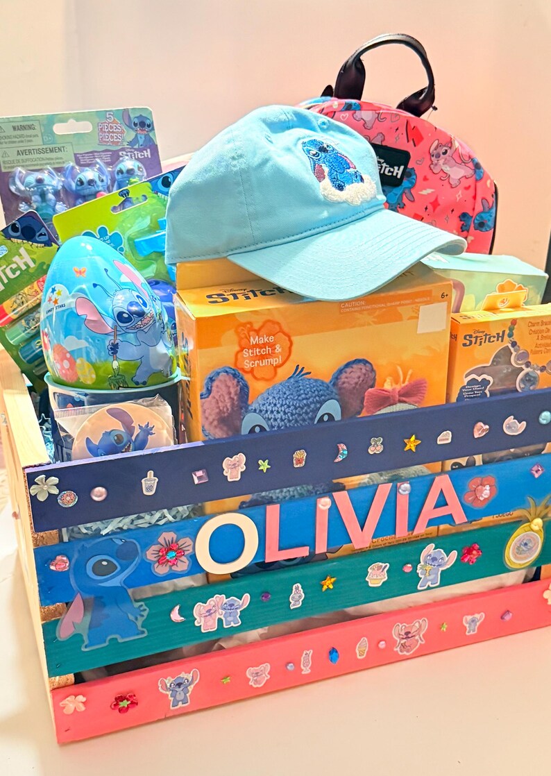 K&ouml;nnte beinhalten: Ein Geschenkkorb voller Stitch-Artikel. Enth&auml;lt eine hellblaue Baseballkappe mit einem gestickten Stitch-Design, einen rosa Rucksack, Spielzeug und eine Holzkiste, die mit dem Namen "OLIVIA" und Stitch-Aufklebern verziert ist.