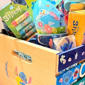 K&ouml;nnte beinhalten: Eine Holzkiste gef&uuml;llt mit Disney Stitch-Merchandise. Zu den Artikeln geh&ouml;ren Lippenbalsam, Figuren, ein Becher und ein eif&ouml;rmiger Beh&auml;lter. Die Kiste ist mit Stitch-Aufklebern und farbenfrohen bemalten Akzenten verziert.