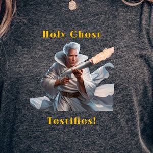 Puede incluir: Una camiseta gris oscuro con un estampado gráfico de un hombre con una túnica blanca sosteniendo una antorcha en llamas. El texto "Holy Ghost Testifies!" está impreso en amarillo debajo del gráfico.