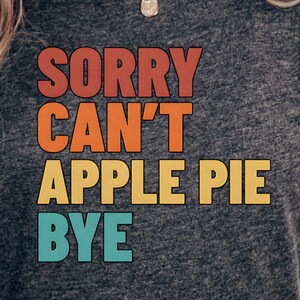 May include: Dark gray t-shirt with the text "SORRY CAN'T APPLE PIE BYE" in retro orange, yellow, and teal lettering. The shirt has short sleeves and a crew neck.