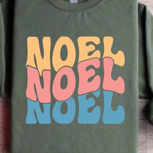 以下が含まれることがあります： レトロなカラフルなフォントで「NOEL」という単語が3回書かれた緑色のスウェットシャツ。スウェットシャツは、松の枝と赤いギフトボックスのある木製の表面に平らに置かれています。