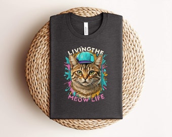 May include: A dark grey t-shirt featuring a graphic of a cat wearing a blue baseball cap and a gold chain. The text "Living the Meow Life" is printed below the cat.