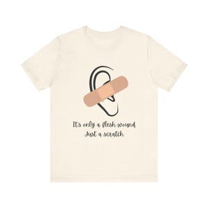 Może przedstawiać: Biały t-shirt z czarno-białą ilustracją ucha z opatrunkiem. Pod ilustracją znajduje się napis "It's only a flesh wound just a scratch".