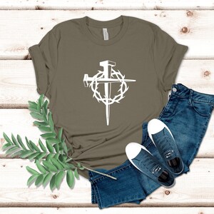 May include: A green t-shirt with a white graphic of a cross with nails and a crown of thorns. The shirt is laid out on a wooden surface with a pair of blue jeans and a pair of white sneakers.