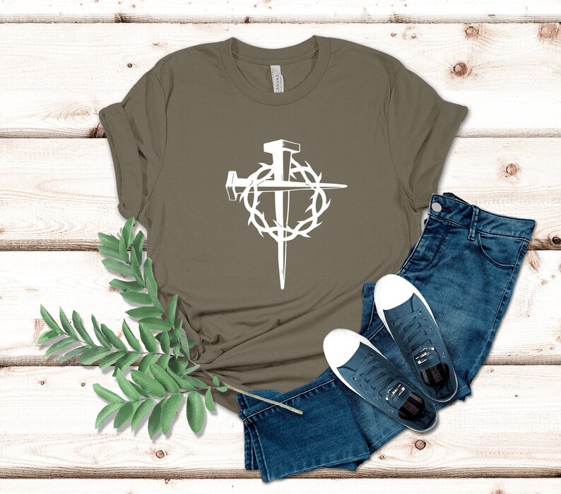 May include: A green t-shirt with a white graphic of a cross with nails and a crown of thorns. The shirt is laid out on a wooden surface with a pair of blue jeans and a pair of white sneakers.