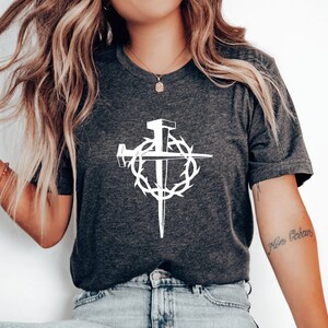 May include: A dark grey t-shirt with a white graphic of a cross with nails and a crown of thorns. The text "Niro Ocean" is tattooed on the wearer's arm.
