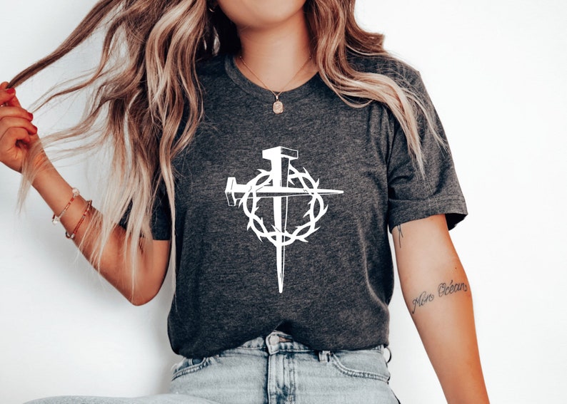 May include: A dark grey t-shirt with a white graphic of a cross with nails and a crown of thorns. The text "Niro Ocean" is tattooed on the wearer's arm.