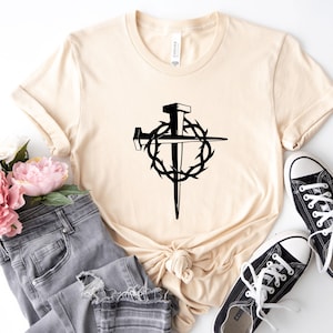 May include: A beige t-shirt with a black graphic of a cross with a crown of thorns and nails. The shirt is laying on a white surface with a pair of black sneakers and a pair of grey jeans.