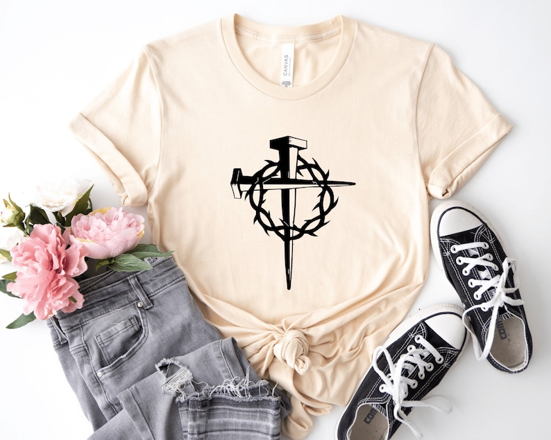 May include: A beige t-shirt with a black graphic of a cross with a crown of thorns and nails. The shirt is laying on a white surface with a pair of black sneakers and a pair of grey jeans.