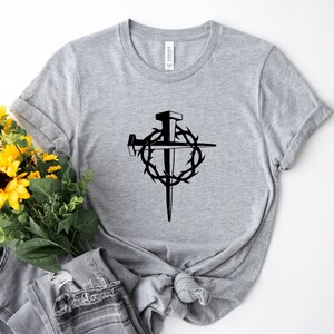 May include: A gray t-shirt with a black graphic of a cross with a crown of thorns and nails. The graphic is centered on the shirt.