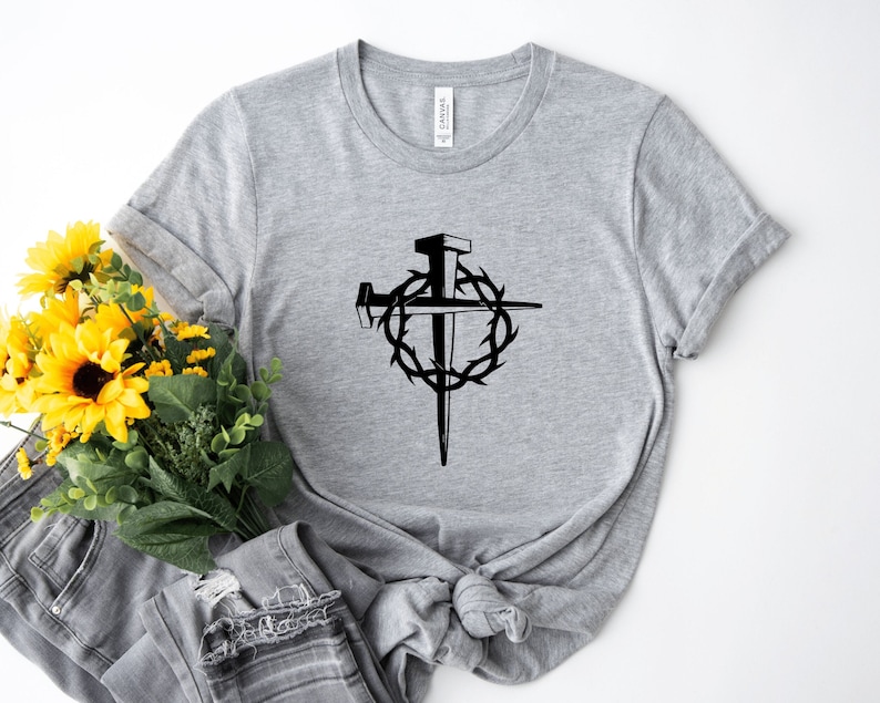 May include: A gray t-shirt with a black graphic of a cross with a crown of thorns and nails. The graphic is centered on the shirt.