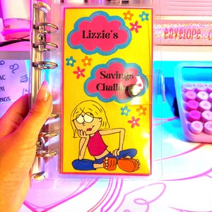 May include: A clear plastic binder with a yellow page insert. The page has a cartoon girl and the text "Lizzie's Savings Challenge".