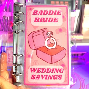 May include: A pink and white planner page with the text "Baddie Bride Wedding Savings". The page features a cartoon illustration of a ring box with a diamond ring inside.