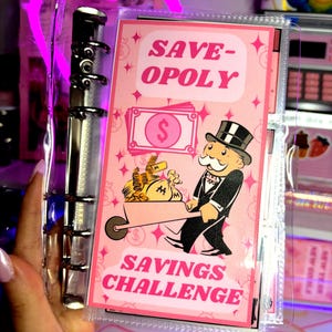 May include: A pink and silver binder with a "Save-Opoly Savings Challenge" insert. The insert features a Monopoly-themed design with a cartoon character pushing a wheelbarrow of money. The binder is held in a person's hand.