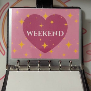May include: A black binder with a pink and yellow heart-shaped sticker that says "WEEKEND" on it. The sticker has a pink background with yellow stars.