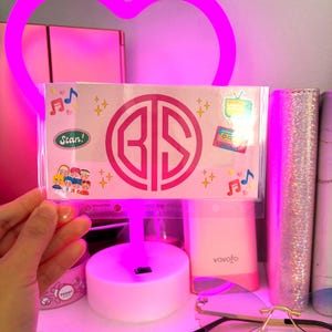 May include: A pink heart-shaped neon light illuminates a white card with the initials "BS" in a circle. The card is decorated with stickers, including a cartoon character, a cassette tape, and a television set. The card is sitting on a white shelf with other items.