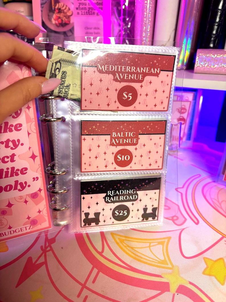 May include: A clear binder with three property cards labeled "Mediterranean Avenue", "Baltic Avenue", and "Reading Railroad". The cards are pink and black with white text and star accents. A hand is holding a five-dollar bill.
