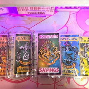May include: Five clear plastic binders with colorful Harry Potter house crests. Each binder has a different house crest: Gryffindor, Slytherin, Hufflepuff, Ravenclaw, and Hogwarts. The binders have the word "SAVINGS" printed on them.