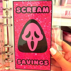 May include: A pink glitter binder with a clear plastic cover. The binder has a black and pink graphic of the Ghostface mask from the Scream movie franchise. The text "SCREAM SAVINGS" is printed on the binder.