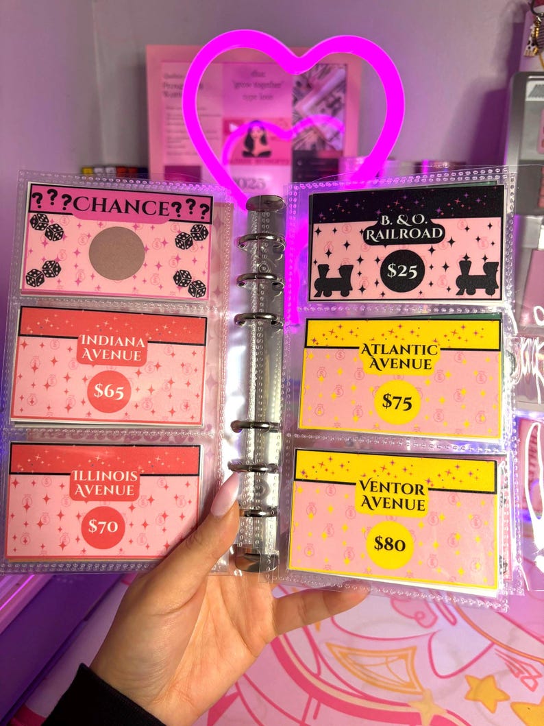 May include: A clear binder displaying Monopoly-themed cards with pink and yellow backgrounds. The cards feature text like "Chance," "Indiana Avenue," and "B. & O. Railroad." A bright pink heart-shaped light is above the binder.