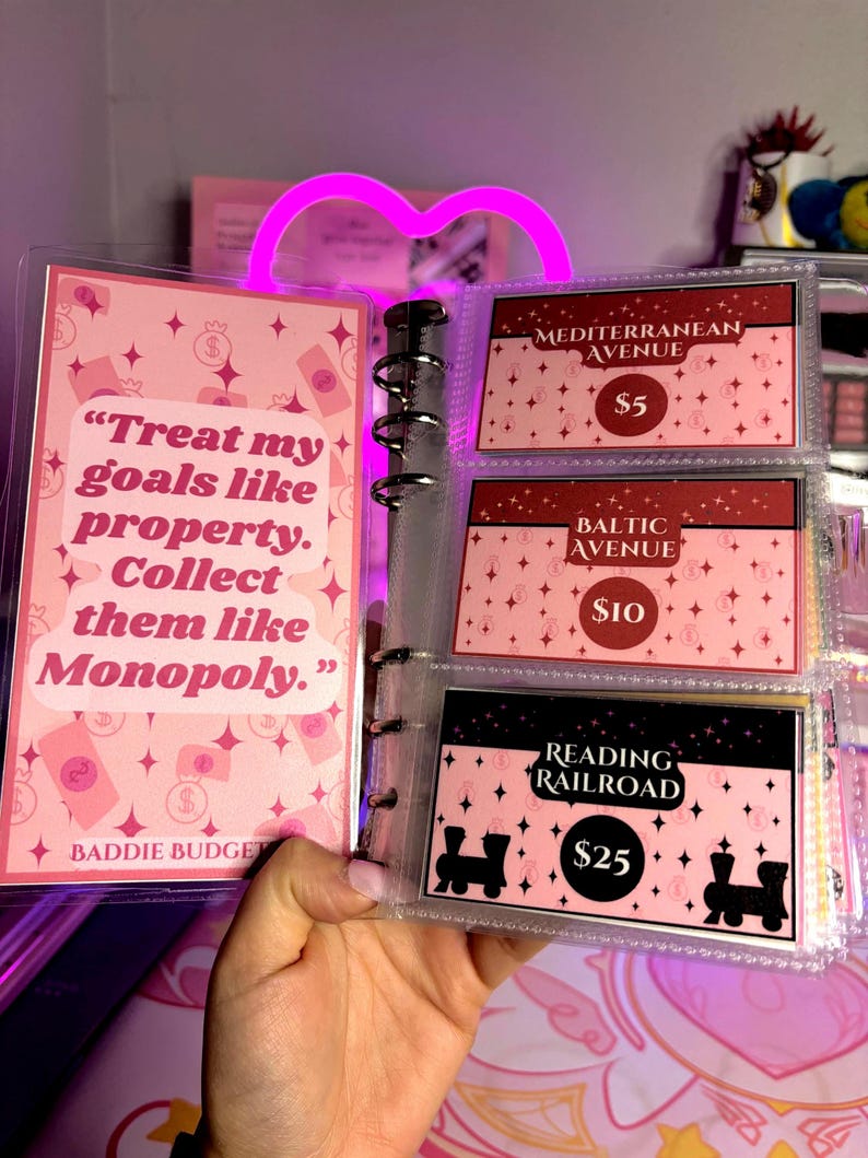 May include: A pink binder with inserts featuring Monopoly-themed financial planning cards. The cards display phrases like "Treat my goals like property" and "Collect them like Monopoly." The binder includes cards for Mediterranean Avenue, Baltic Avenue, and Reading Railroad.