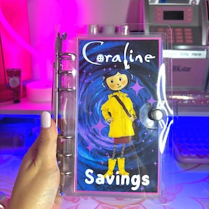 May include: A clear plastic binder with a colorful insert featuring Coraline from the movie Coraline. The insert has a swirling blue and purple background with stars and the text "Coraline Savings".
