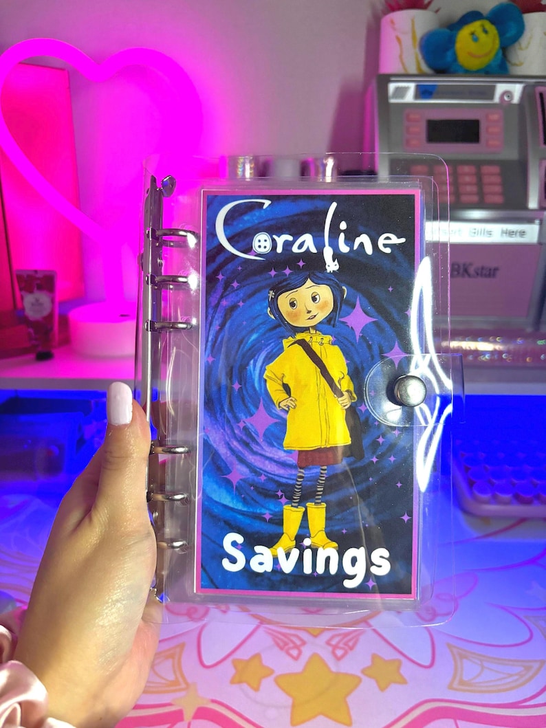 May include: A clear plastic binder with a colorful insert featuring Coraline from the movie Coraline. The insert has a swirling blue and purple background with stars and the text "Coraline Savings".