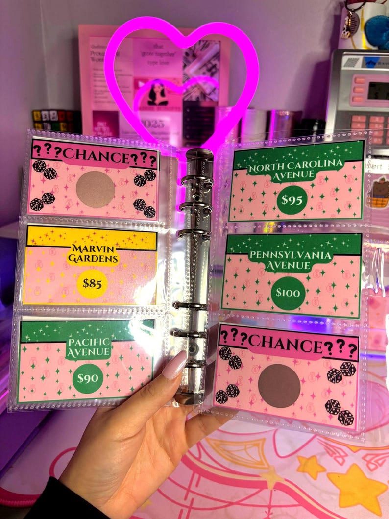 May include: A clear binder with pink and green cards featuring text like "Chance", "Marvin Gardens", and "Pacific Avenue". The cards have a pink background with a diamond pattern. A neon pink heart-shaped light is attached to the binder.