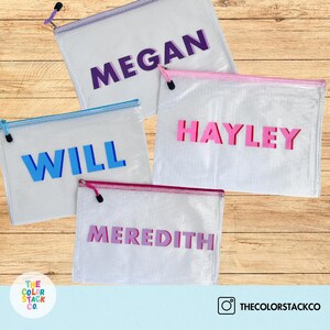 May include: Four clear mesh zippered pouches with different colored names on them. The names are Megan, Will, Hayley, and Meredith. The pouches are on a wooden surface.