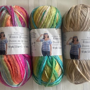 May include: Three skeins of Ribbon Dancer yarn in vibrant colors. One skein is a mix of pink, orange, and purple. Another is green, yellow, and blue. The third is a neutral tan color. Each skein has a label with a woman wearing a hat and the text "Ribbon Dancer".