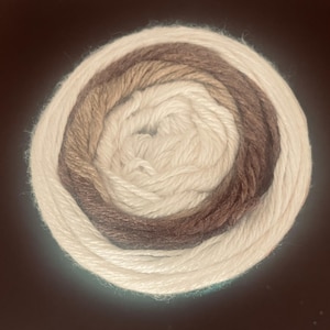 May include: A tightly wound ball of yarn in a gradient of neutral colors. The yarn transitions from a creamy white to light brown, medium brown, and then a darker brown at the center. The yarn is likely for knitting or crocheting projects.