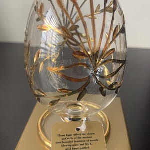 Könnte beinhalten: Ein klarer, eiförmiger Glasornament mit Goldblattakzenten, der auf einem goldfarbenen Ständer ruht. Das Ornament hat ein Blumendesign und ein Etikett mit der Aufschrift "These Eggs reflect the charm and style of the ancient time honored tradition of mouth blowing glass and 24 K. gold hand painted Made in Egypt."