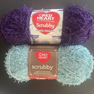 Puede incluir: Dos ovillos de hilo Red Heart Scrubby, uno morado y otro azul claro. El ovillo morado está arriba, el azul claro abajo. El embalaje dice "Great for Dishcloths!" Ideal para paños de cocina.