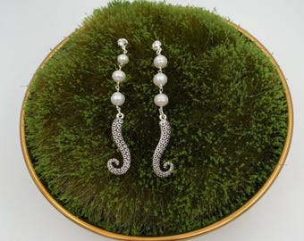 May include: A pair of silver dangle earrings with a pearl and octopus design. The earrings are on a green mossy surface.