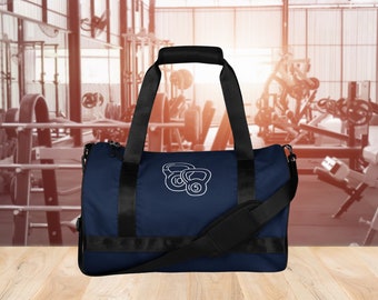 May include: A navy blue duffel bag with black straps and a white graphic of three kettlebells. The bag is sitting on a wooden surface.