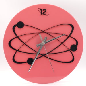 May include: A pink round clock with black hands and a black atomic symbol design. The clock face has the number 12 at the top.