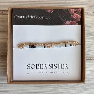 May include: A beaded bracelet with the words "SOBER SISTER" on a white card, presented in a brown box. The bracelet features gold, black, and light blue beads. The card also includes Morse code.