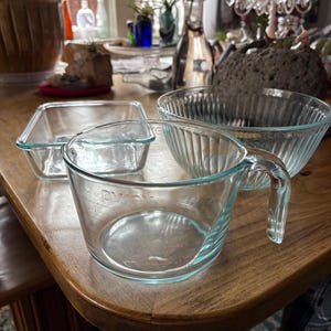 May include: A collection of clear glass Pyrex measuring cups and bowls on a wooden table. The measuring cup has a handle and measurement markings. A square baking dish and a round bowl are also visible.