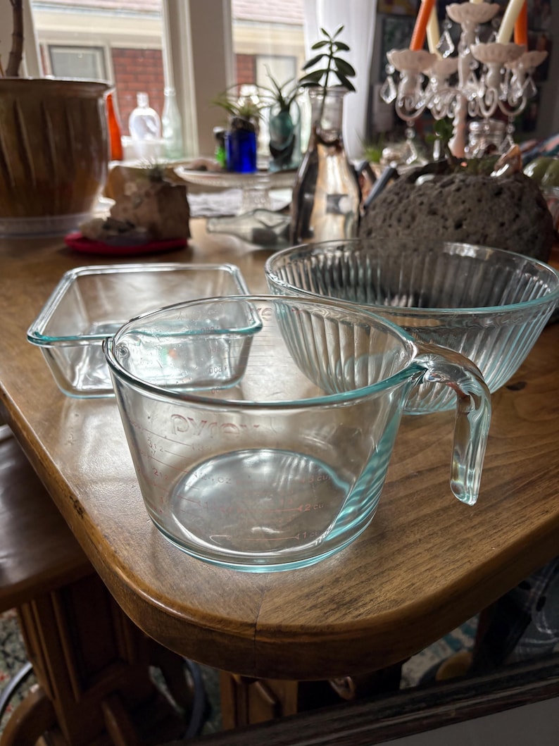 May include: A collection of clear glass Pyrex measuring cups and bowls on a wooden table. The measuring cup has a handle and measurement markings. A square baking dish and a round bowl are also visible.