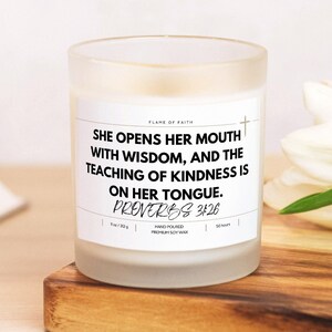 May include: A frosted glass candle with a white label featuring the text "She opens her mouth with wisdom, and the teaching of kindness is on her tongue. Proverbs 3:26." The candle is hand-poured with premium soy wax and has a burn time of 50 hours.