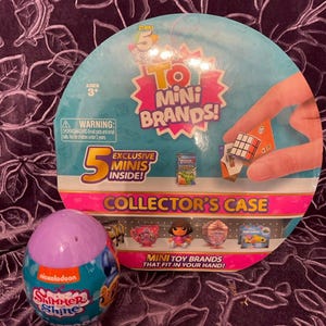 May include: A purple and blue egg-shaped container with the Nickelodeon logo and the words "Shimmer Shine" on it. The container is open and shows a small plastic toy inside. The container is sitting on a purple background with a larger round container behind it. The larger container is pink and white and has the words "5 To Mini Brands!" on it. The larger container also has a picture of a Rubik's Cube and the words "Exclusive Minis Inside!" on it.