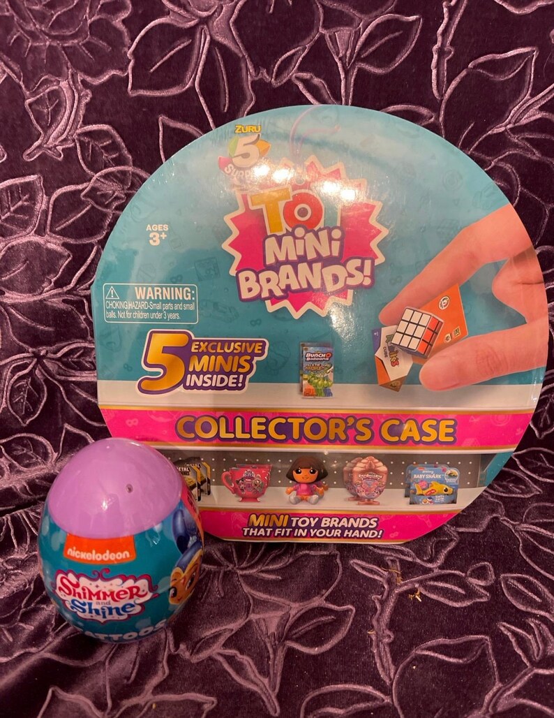 May include: A purple and blue egg-shaped container with the Nickelodeon logo and the words "Shimmer Shine" on it. The container is open and shows a small plastic toy inside. The container is sitting on a purple background with a larger round container behind it. The larger container is pink and white and has the words "5 To Mini Brands!" on it. The larger container also has a picture of a Rubik's Cube and the words "Exclusive Minis Inside!" on it.