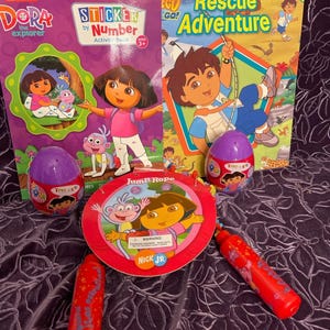 May include: Dora the Explorer themed children's books and toys.  The books include "Sticker by Number Activity Book" and "Animal Rescue Adventure".  The toys include a jump rope, two egg-shaped toys, and a purple and red toy.