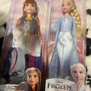 May include: Two Disney Frozen dolls, Anna and Elsa, in their original packaging. Anna wears a purple and black dress with a brown belt. Elsa is in a light blue dress with a white bodice. The packaging features the Frozen logo and character images.