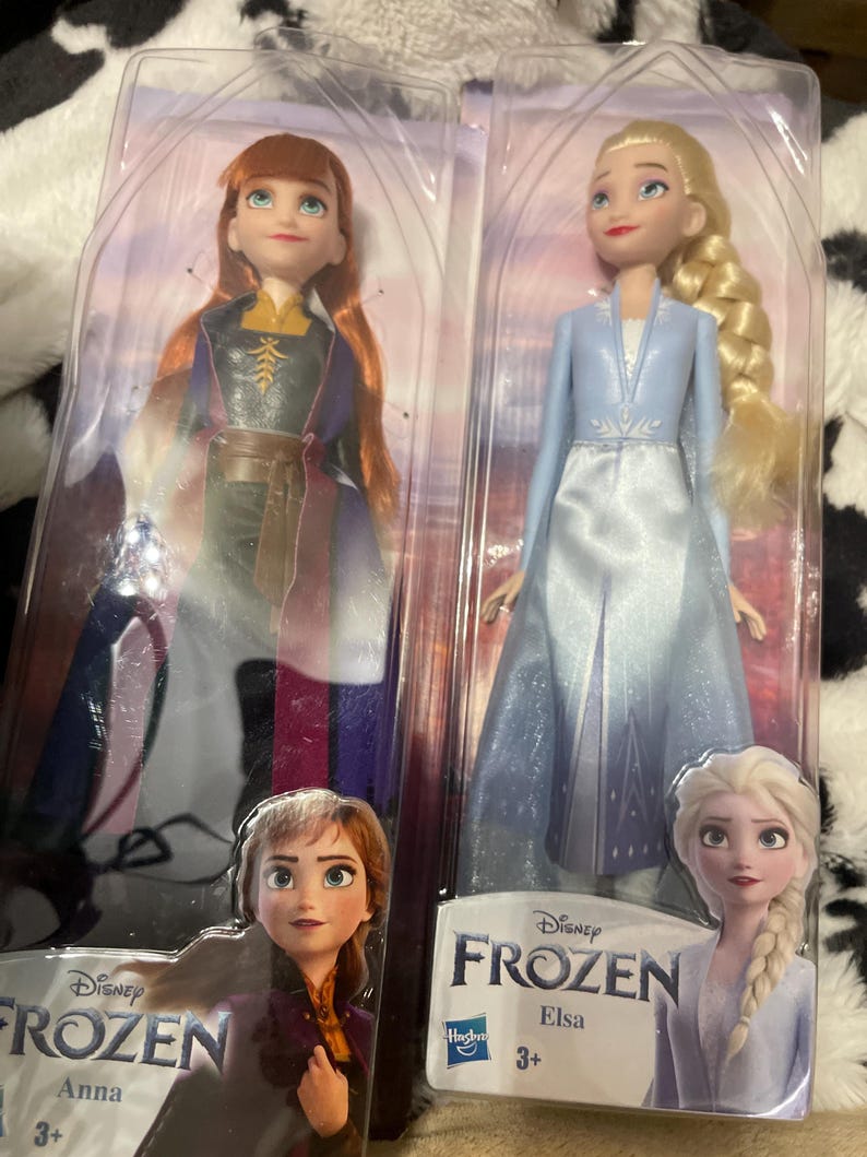 May include: Two Disney Frozen dolls, Anna and Elsa, in their original packaging. Anna wears a purple and black dress with a brown belt. Elsa is in a light blue dress with a white bodice. The packaging features the Frozen logo and character images.