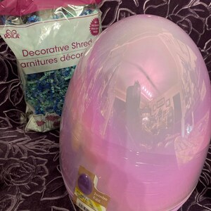 May include: A large iridescent pink plastic egg with a label that reads "Jumbo Egg". The egg is sitting on a purple fabric surface.